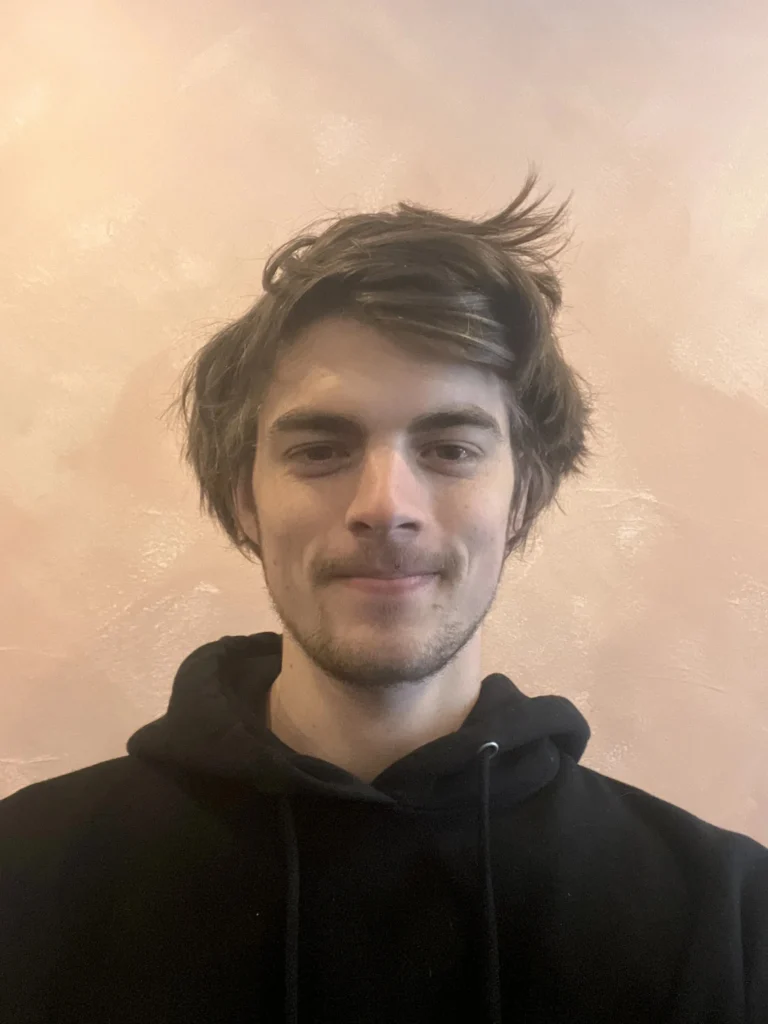 Cassius Luber, holistic health and movement coach, smiling in a black hoodie against a soft pink background, reflecting a welcoming and approachable demeanor.