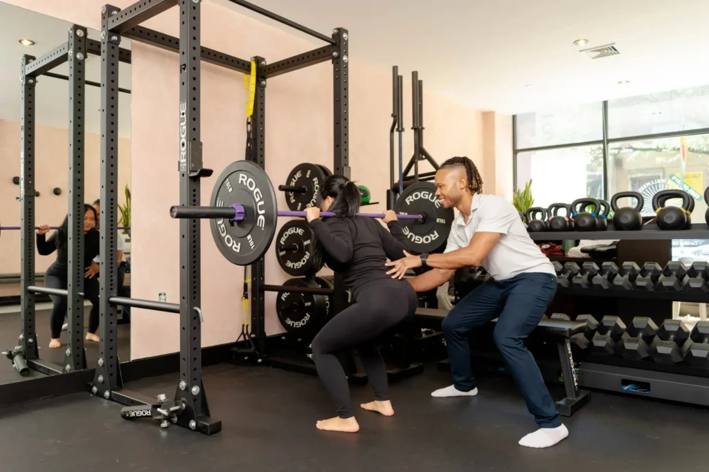 Woman performing a squat with a barbell under the guidance of a trainer in a fitness studio, emphasizing functional fitness and strength training for improved mobility.