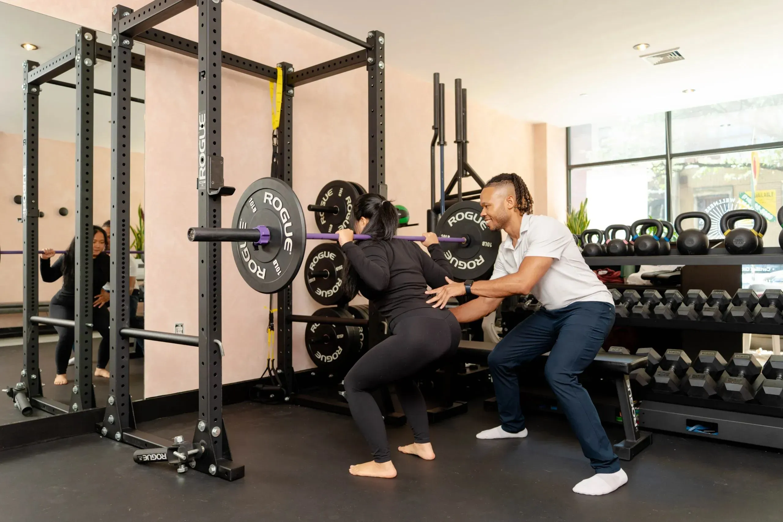 Personal Training Hunters Point: Tailored Fitness Programs 1 Personal trainer assisting client with barbell squat in Dekhana Wellness studio in Hunters Point.